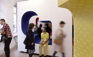 View of a white and blue installation which has a man dressed in yellow sitting inside it talking to a woman who is standing up. Through the centre of the installation you can see another white and pink installation and several other people