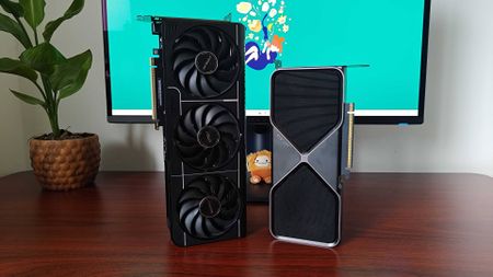 RTX 5070 Founders Edition and Asus Prime OC graphics card standing vertical on woodgrain desk next to plant and monitor