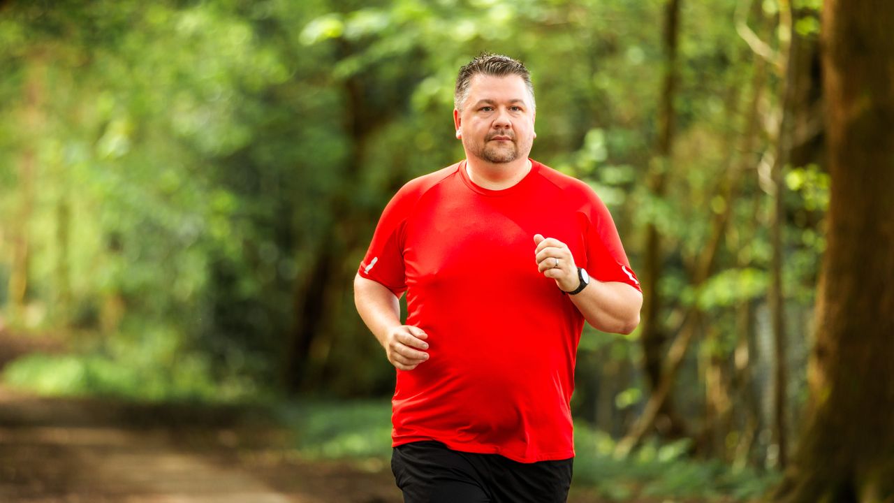 A man in a bright red t-shirt runs along a dirt path. Behind him we see blurred trees.