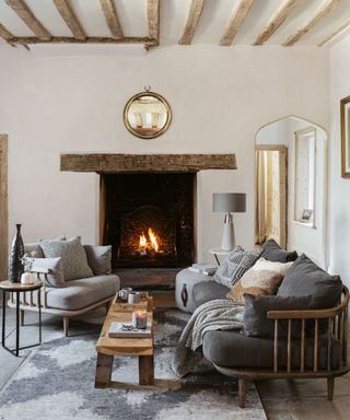A cozy living room with a long wooden sofa and a matching smaller chair on a grey rug next to a roaring fireplace with wooden beam mantle