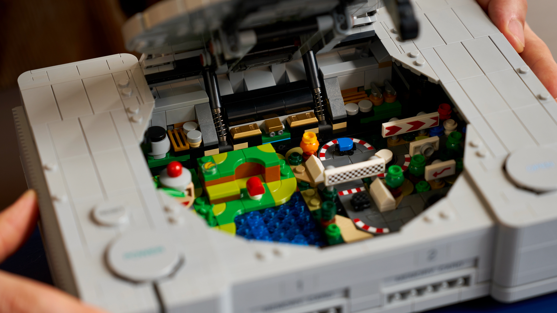 Hands hold an open Lego PlayStation model, with the miniature dioramas inside visible