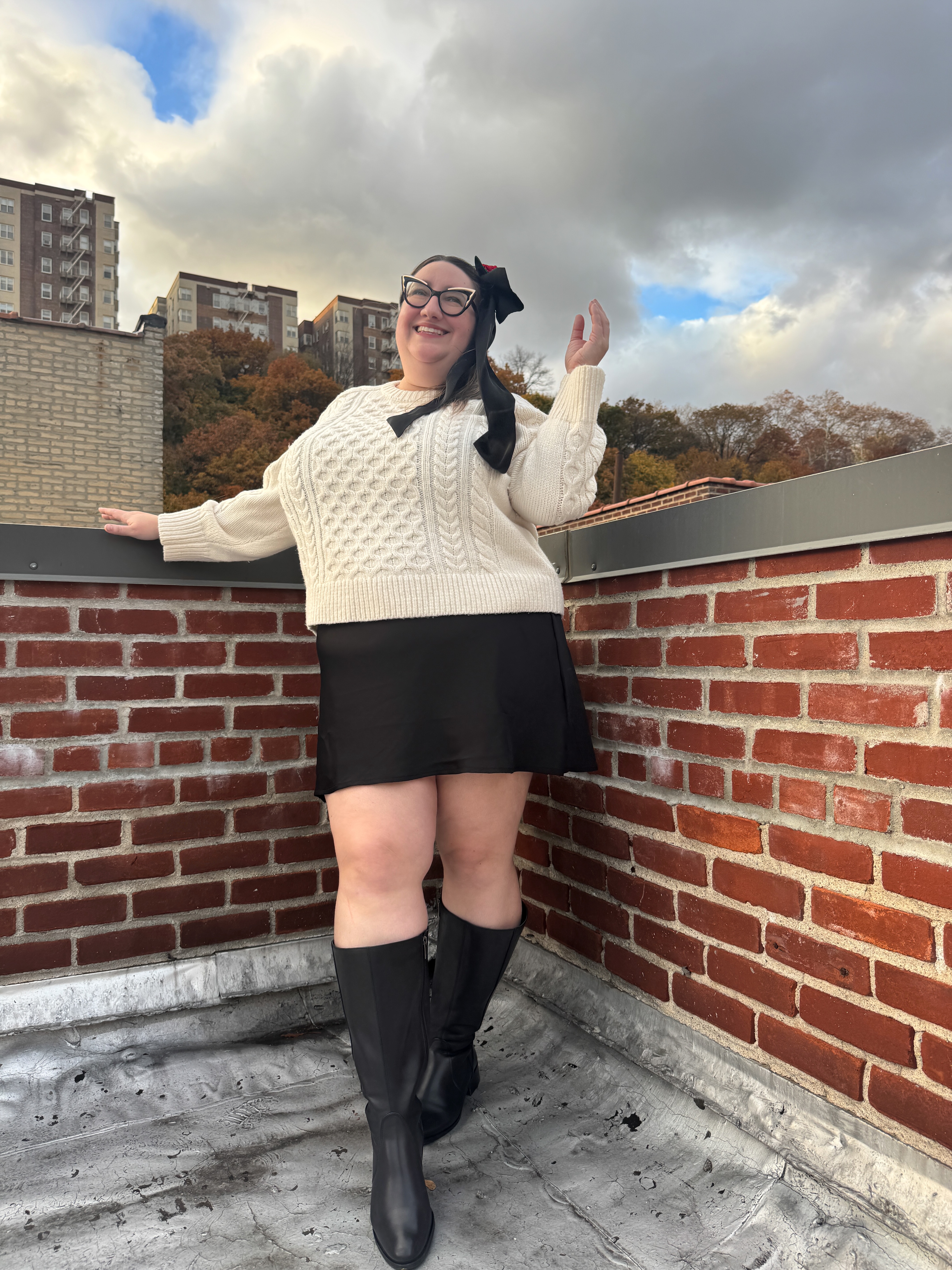 Sarah Chiwaya wearing a little black dress, cream cable knit sweater, and tall leather boots