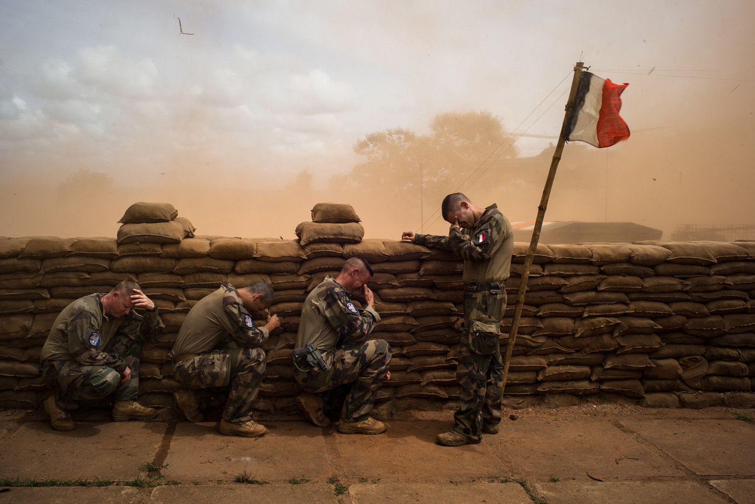 four soldiers crouching behind sandbags with a french flag, there is lots of dessert dust in the air