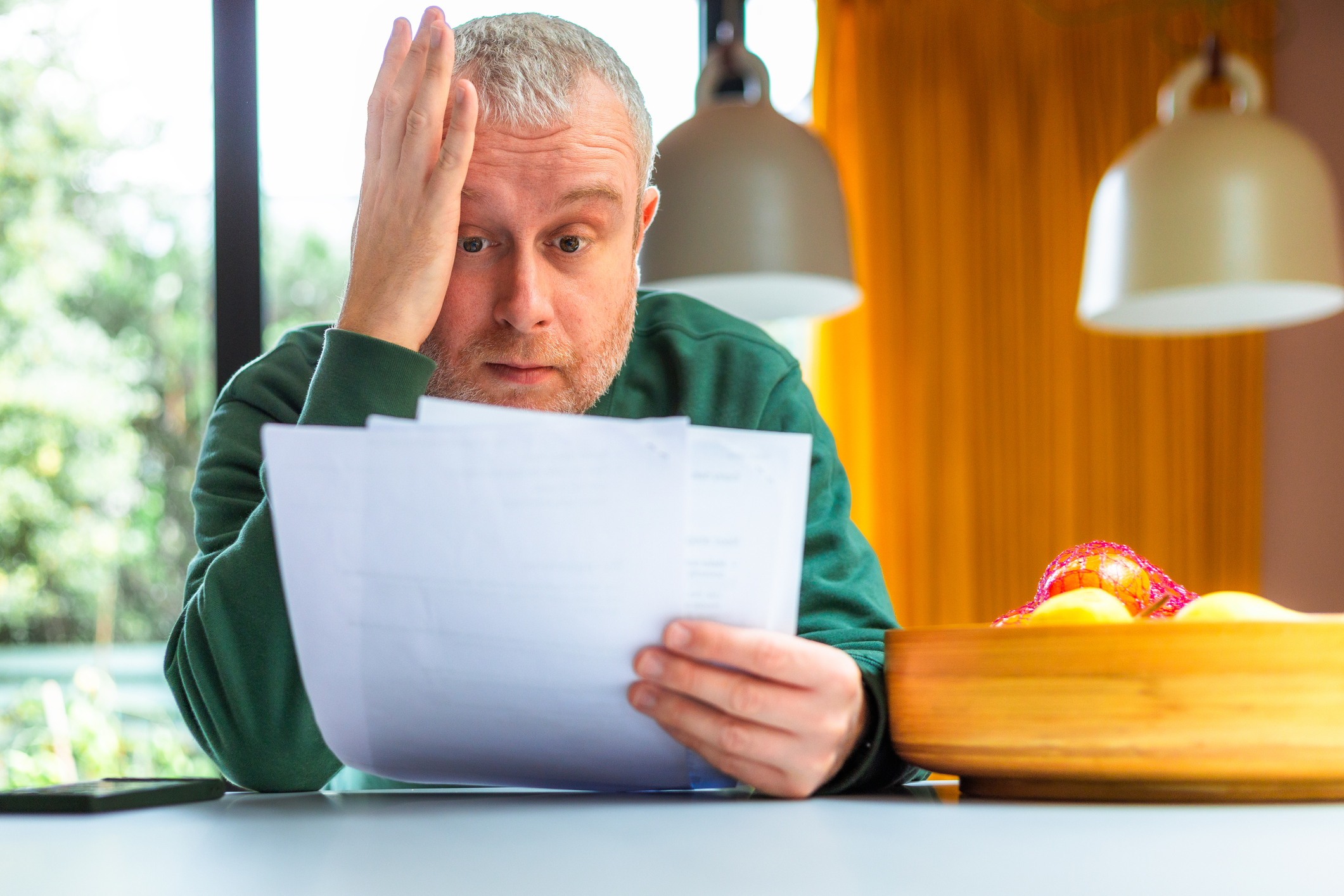 Portrait of a mid adult man checking his energy bills at home. He has a worried expression and touches his face with his hand while looking at the bills. He is sitting in his modern kitchen at home, dressed casually in green crewneck sweater and blue denim jeans.