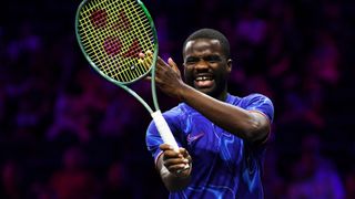 Frances Tiafoe laughs as he practices at the 2024 Laver Cup wearing a blue t-shirt and holding his racket aloft.