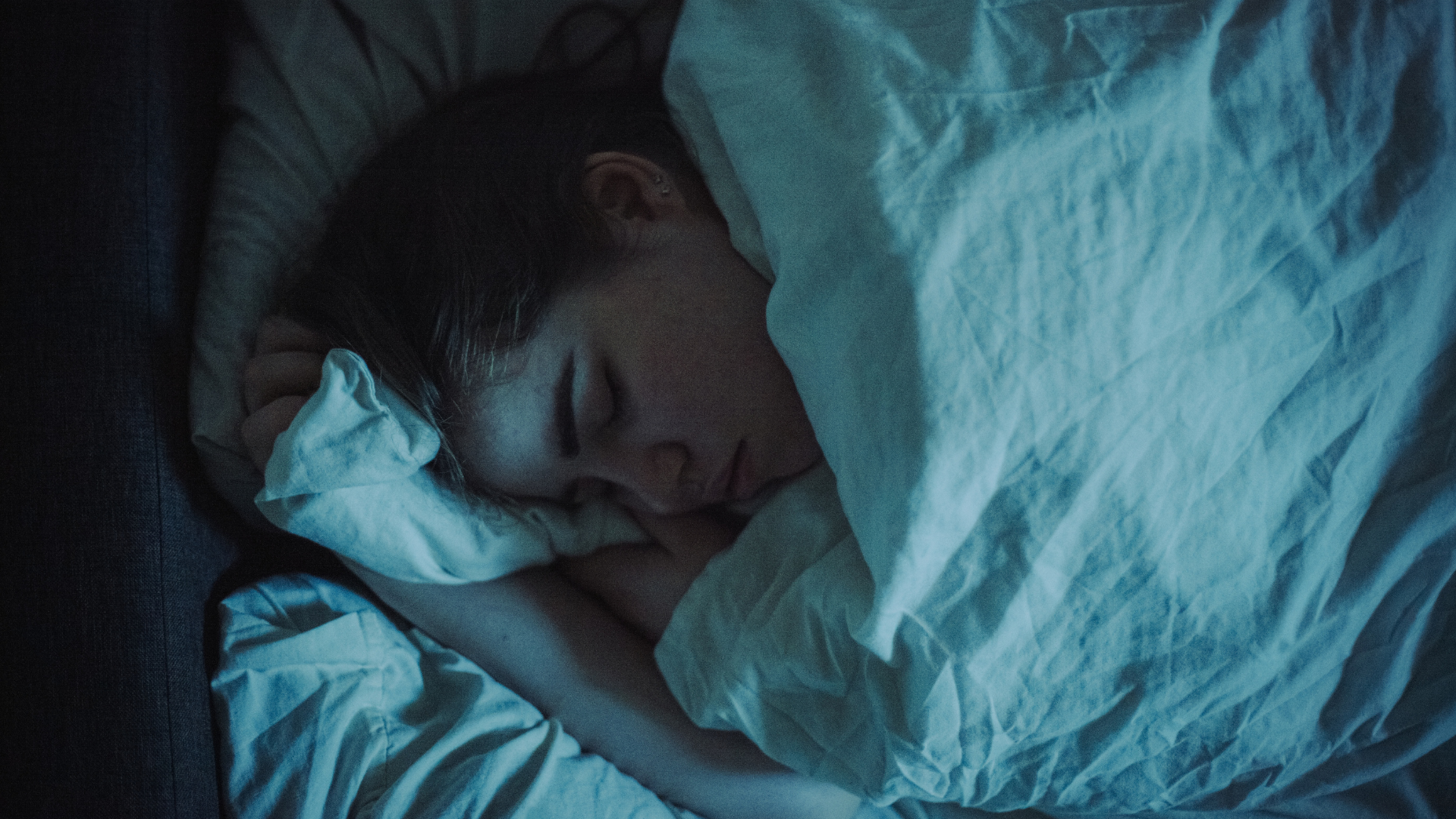 A woman asleep on her side in a bed at night, the bed has white bedding.