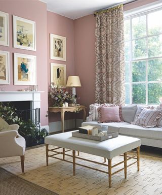 A vintage style pink living room with patterned curtains in front of a large window, with white framed gallery wall, a white mantelpiece, a grey sofa with frilly pink and striped cushions with matching footrest with tray and a blush pink armchair facing towards it