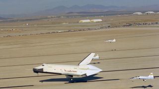 space shuttle touching down on a desert runway with three planes landing around it. mountains and buildings are in the background