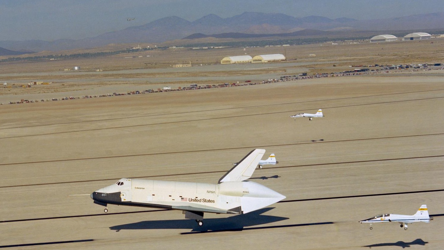 space shuttle touching down on a desert runway with three planes landing around it. mountains and buildings are in the background