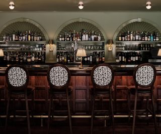 green hotel bar with a large dark wood panelled bar, upholstered wooden stools, and arched scalloped shelving filled with drinks