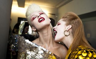 Two female models backstage, in designer outfits, bright makeup, one model tilting her head back with eyes closed the other jokingly pretending to bite her neck, white walls, wall lights shining