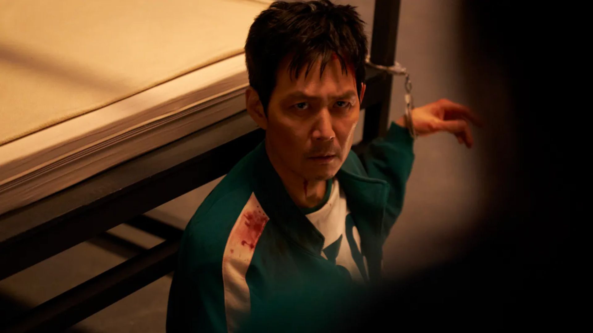 an image of Lee Jung-jae playing as Gi-hun in squid game season three, handcuffed to a railing in a green tracksuit