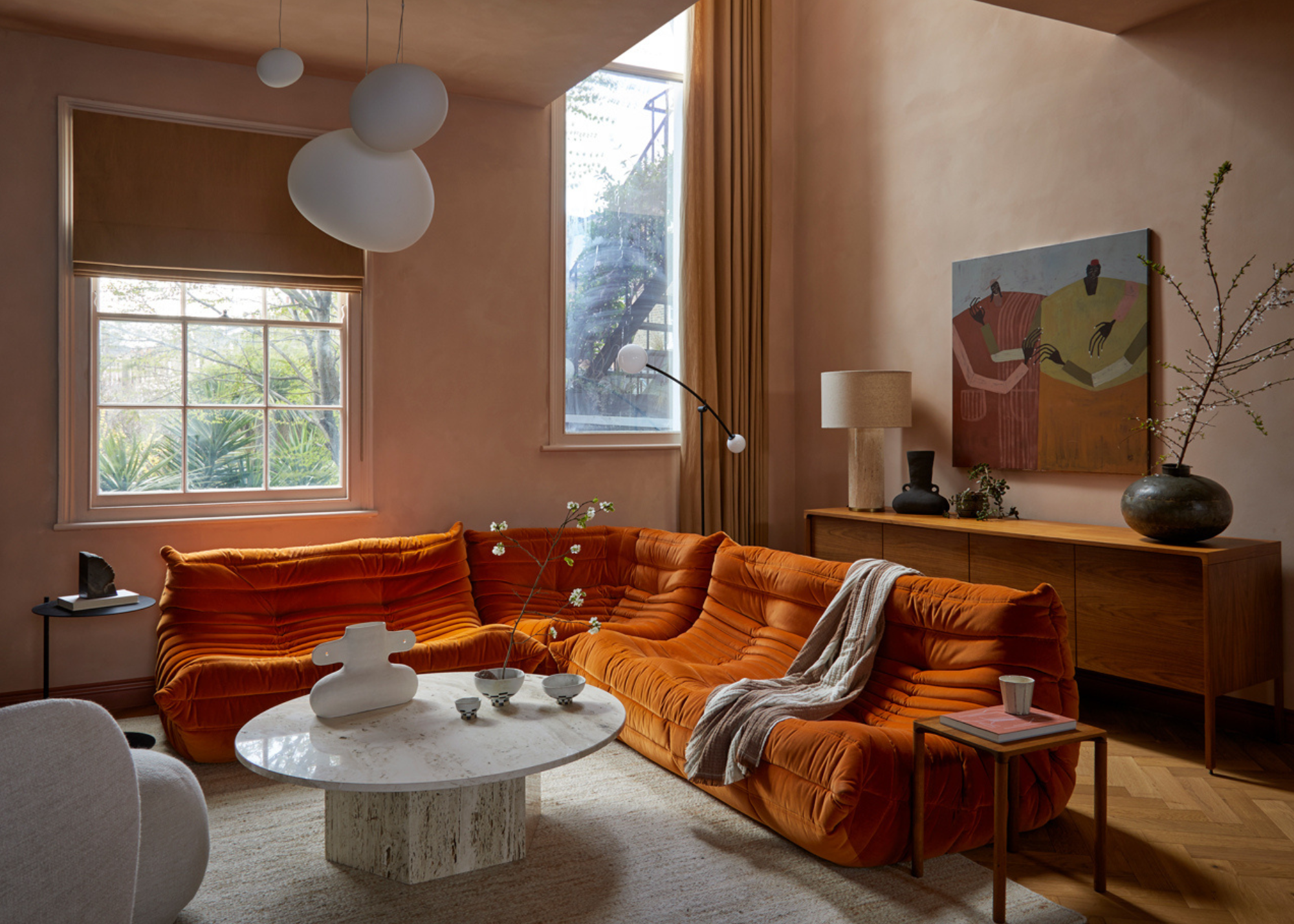 A bright living room with beige pink walls, a large painting, a wood sideboard, orange togos, a stone coffee table, a cream rug, a vase, and table lamp