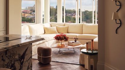 Image of a living room in a sun room nook off a kitchen that has a marble island in view. The nook has a large cream sectional and a small coffee table.