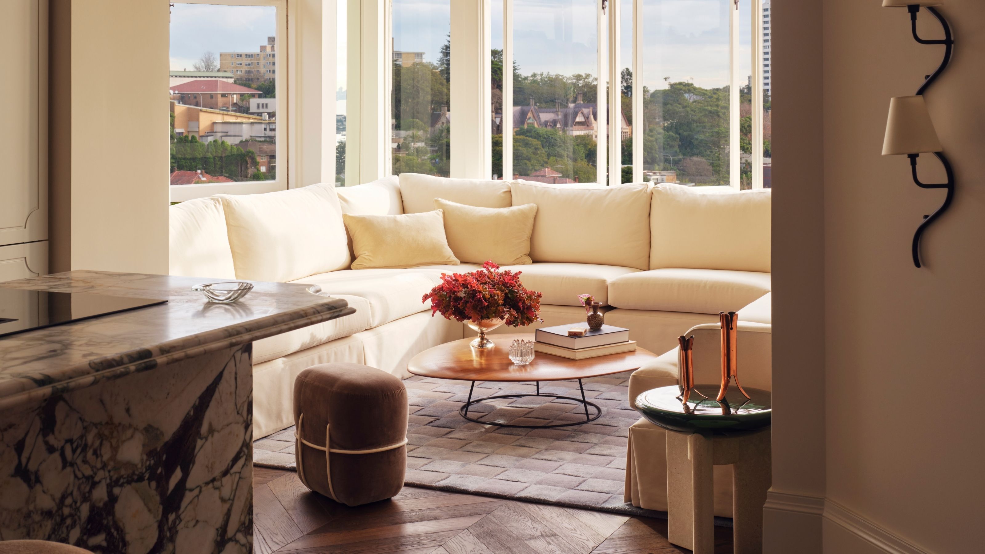 Image of a living room in a sun room nook off a kitchen that has a marble island in view. The nook has a large cream sectional and a small coffee table.