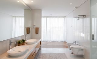 Bathroom featuring white walls and ceiling with brown cabinets and two white sinks with taps below a large wall mirror. His and hers white toilets