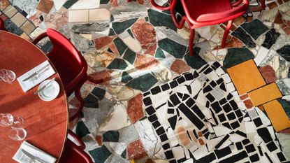 A terrazzo floor made with a speckled surface of uneven fragments of stone in green, terracotta, white, black, beige, and yellow, topped by red-tinted sculptural furniture and captured from above.