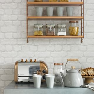 Grey worktop in a kitchen with a white brick wall, with shelves for mugs and small appliances on the worksurface below