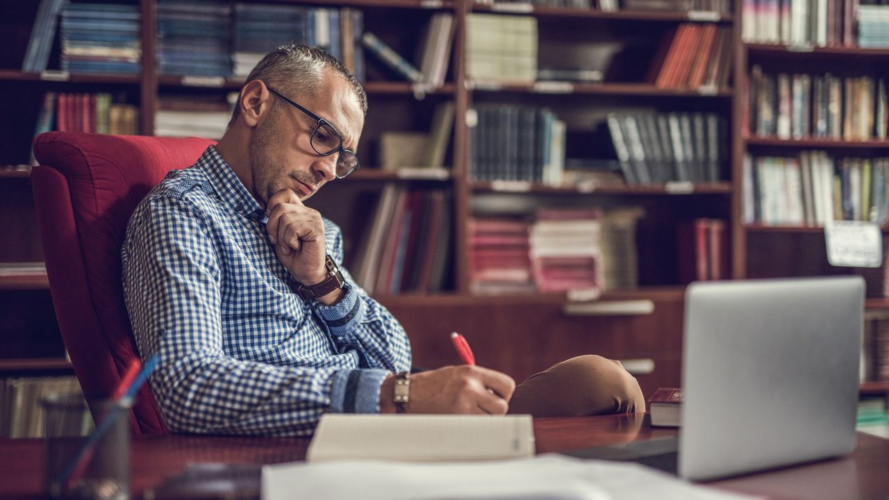 An older business executive sits at his desk and takes notes in front of his laptop.