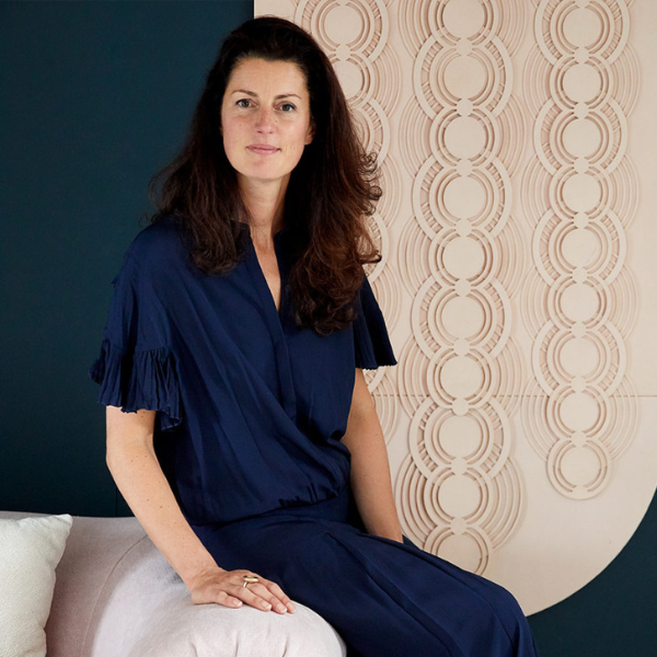 a headshot of genevieve bennett, a woman with lond dark brown hair looking at the camera, sitting on the edge of a sofa wearing a dark blue outfit