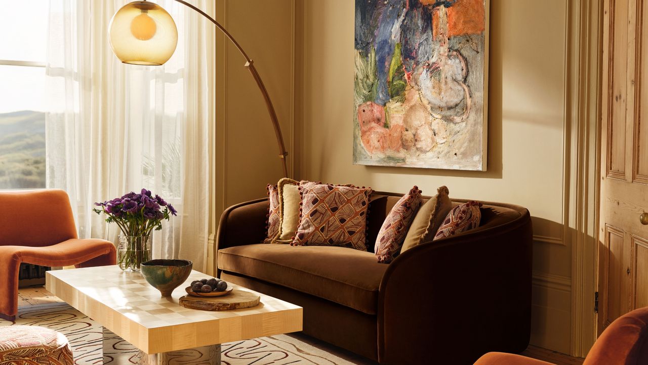 Image of a beige living room with a chocolate brown sofa against the wall. In front of the sofa is a coffee table with a chrome stand and a light wooden top. There are two mid-century-style, burnt orange accent chairs and an arched floor lamp in the corner that has a bulb shade.