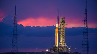 an orange-and-white rocket stands upright under a purple and pink sunset