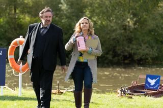 Frank Hathaway (Mark Benton) and Lu Shakespeare (Jo Joyner) walk towards the camera with a boating lack in the background behind them. Lu is carrying a case file and a large reusable pink plastic bottle.