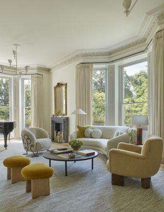 Image of a modern art nouveau living room. There are two large bay windows in the room with elegant, off-white curtains. There is a white curved sofa, a boucle white accent chair, a yellow accent chair, and two yellow stools. There is a round coffee table in the middle of the sitting area.