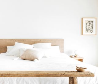 white bedroom with white sheets, timber bedframe, vintage timber bench at the bottom of bed, small timber nightstand with light and mug on it, and an artwork on the wall