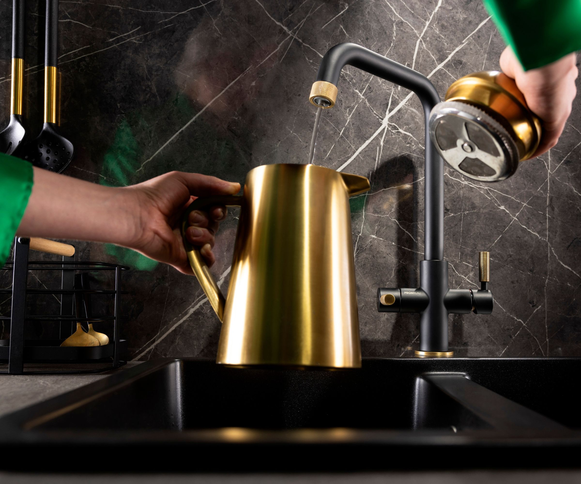 A hand fills a brushed brass coffee pot at a matte black kitchen sink, with a black and gold tap set against a dark marble splashback for a modern, luxury coffee station look.