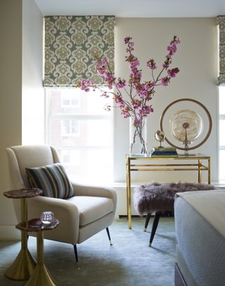 apartment style bedroom with glass and gold vanity, ornaments and books on top with large vase of flowers with blossom in. Armchair, side tables, bed to one side, fluffy sheepskin footstool, patterned blinds