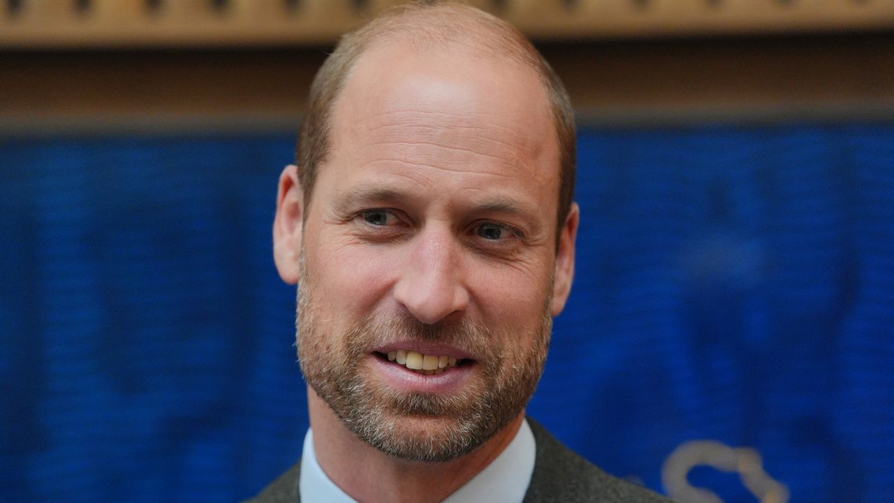 Prince William wearing a gray suit and tie smiling in front of a blue background