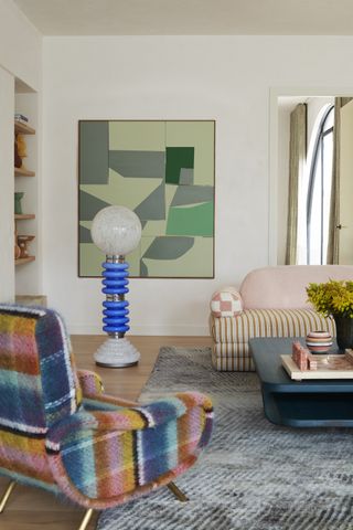 Image of a white living room with a light blue rug. There is a green abstract painting hanging on the wall. There is a light pink and multipatterned cloud couch with a futuristic, electric blue bulb floor lamp beside it. In the foreground, there is a multi-colored plaid accent chair.