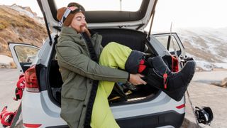 Young man sitting in car trunk putting on snowboard boots