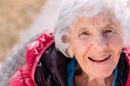 Beautiful, Confident, Sharp 100-Year-Old Elderly Senior Caucasian Woman Wearing a Pink Puffy Jacket Sitting Outdoors in Shade in the the Winter