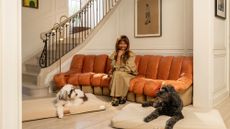 Brigette Romanek sitting on an orange couch in a grand entryway. There is a white dog and a black dog both lay on her designer dog beds