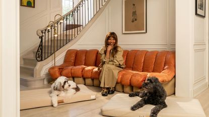 Brigette Romanek sitting on an orange couch in a grand entryway. There is a white dog and a black dog both lay on her designer dog beds