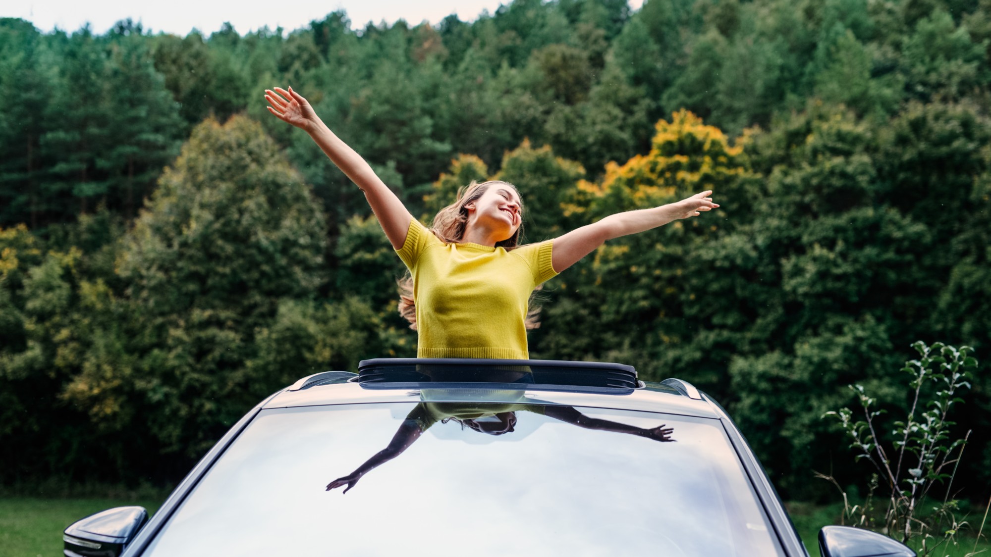 Young woman looking out of car sunroof with arms wide open.