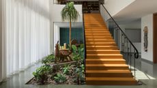a large timber staircase in centre of home lobby with lush garden at its base, sheer curtains, polished concrete floor, and library upstairs
