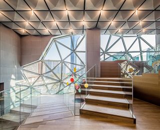 A staircase with wooden floors and glass railings in front of a wall made from different sized triangular windows.