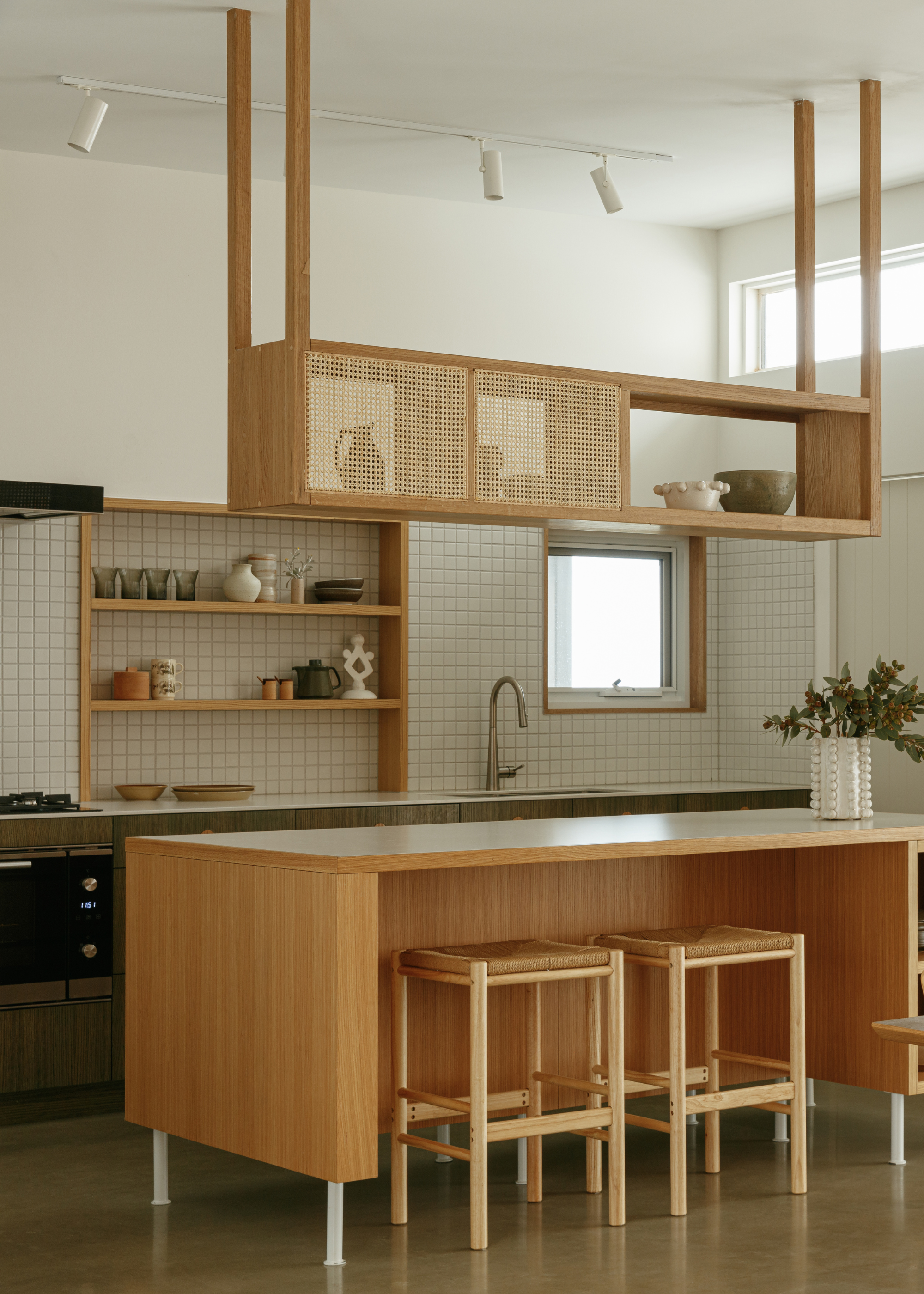 A kitchen with a white tiled backsplash, warm wood shelving, a suspended rattan shelf, a wood island, and a pair of stools