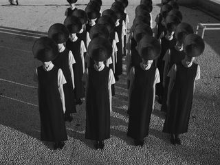 Black and white photo of multiple people standing in rows wearing black and white outfits and black circular hats