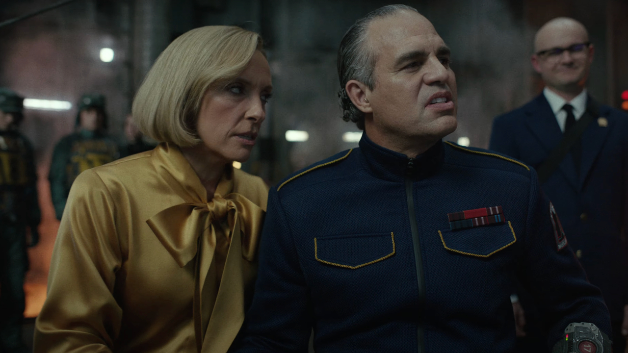 Toni Collette leans to talk to a snorting, uniformed Mark Ruffalo in Mickey 17.