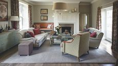 A cozy living room with large rug, upholstered chairs and matching sofas, a glass coffee table, and a large white panelled fireplace. To the left is a blue sideboard. There are bright windows either side with floral curtains.