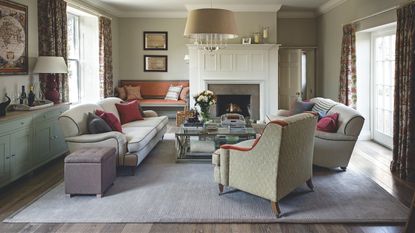 A cozy living room with large rug, upholstered chairs and matching sofas, a glass coffee table, and a large white panelled fireplace. To the left is a blue sideboard. There are bright windows either side with floral curtains.