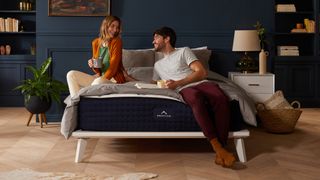 The DreamCloud Mattress photographed on a wooden bedframe placed in a dark blue bedroom and with a couple laughing and sitting on the mattress