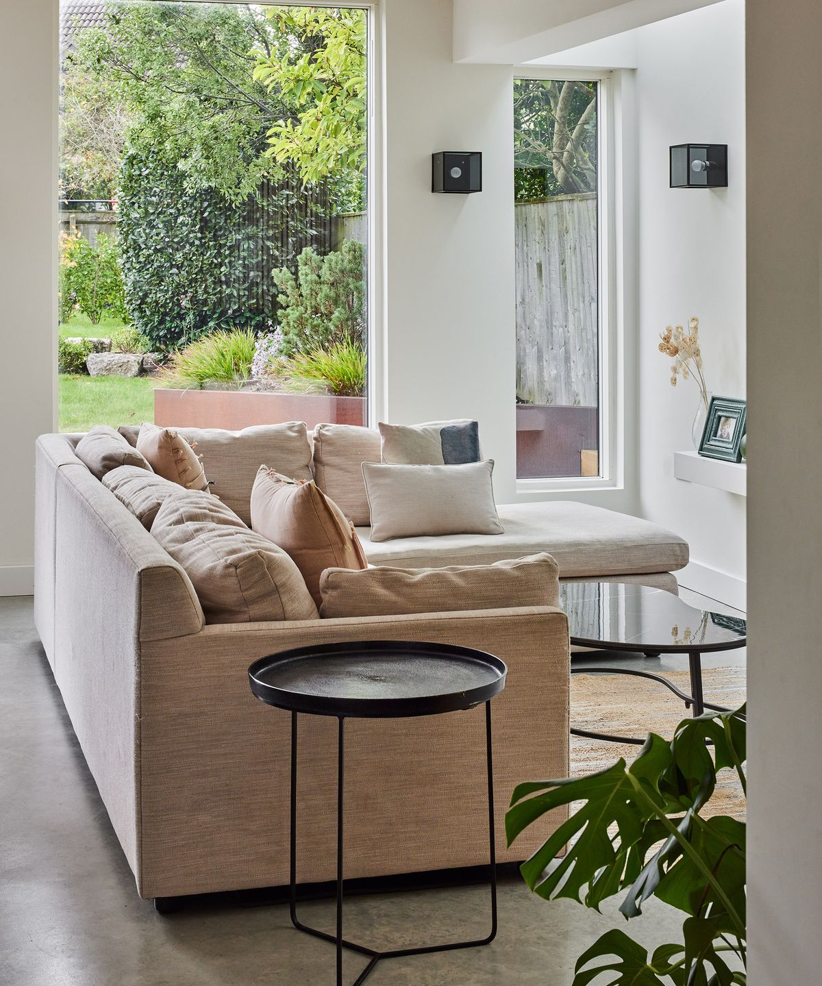 A comfy corner sofa in a light fabric. This lounge nook sits within a large open plan kitchen, diner. The large glazed windows let in copious natural light and have views out into the garden