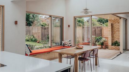 A modern kitchen extension with white floors and walls and countertops, with a small wood table in the center next to a window seat and sliding doors leading to a patio area