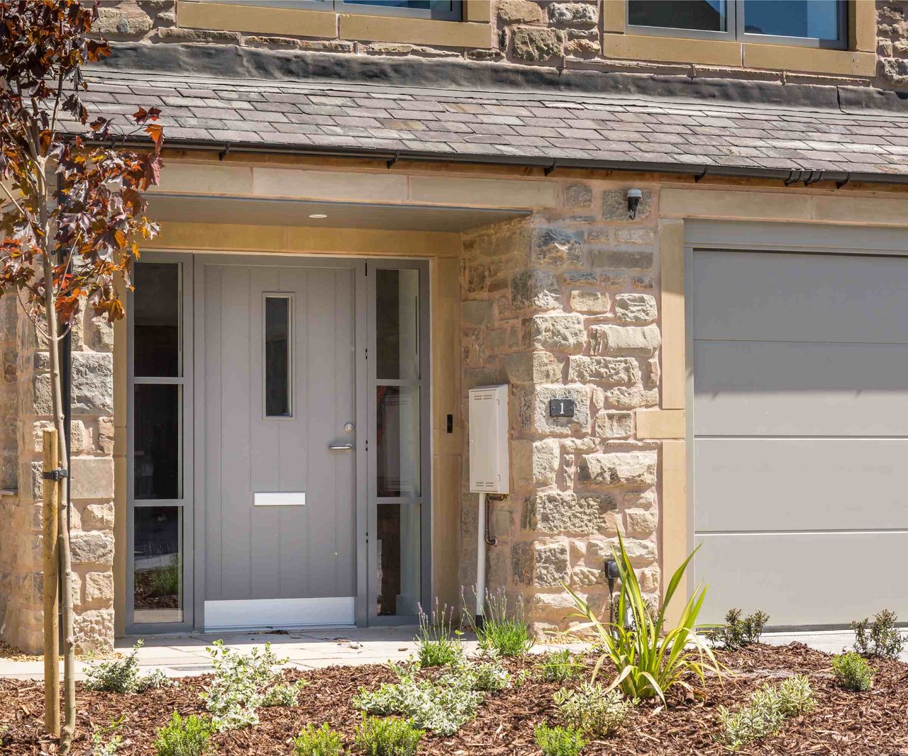 This close-in shot shows part of the front elevation of a light-coloured stone-clad home. The house features a recessed front door, from VELFAC, in a buff, neutral colour, with glazing to either side. The garage door adjacent has been coloured matched to the front door.