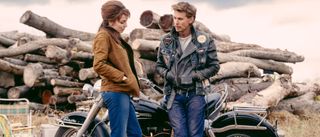 Jodie Comer and Austin Butler stand together talking in front of a bike in The Bikeriders.